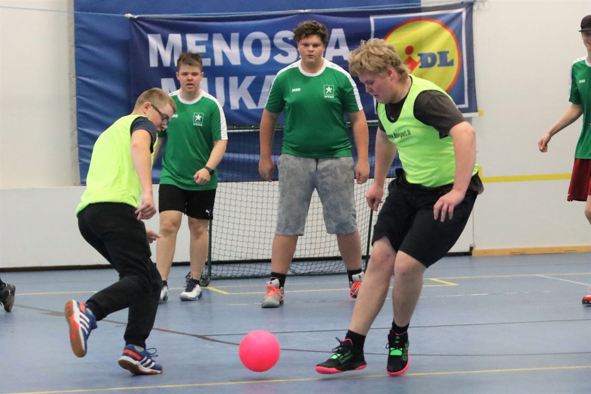 Neljä henkilöä pelaa sisätiloissa jaloillaan palloa. Kahdella päällä vihreät paidat, kahdella vihreät liivit.